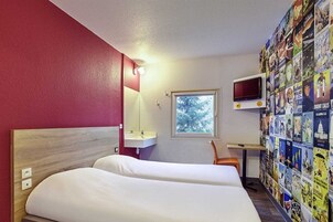 Quarto de hotel com cama, escrivaninha com computador, janela e parede decorada com cartazes.