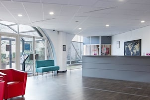 Un área de recepción moderna con entrada por puerta de vidrio, un banco color turquesa y un mostrador de recepción con carteles informativos.