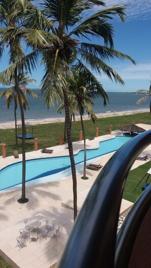 Un área de piscina con palmeras, vista a la playa y un espacio de descanso con mobiliario exterior.