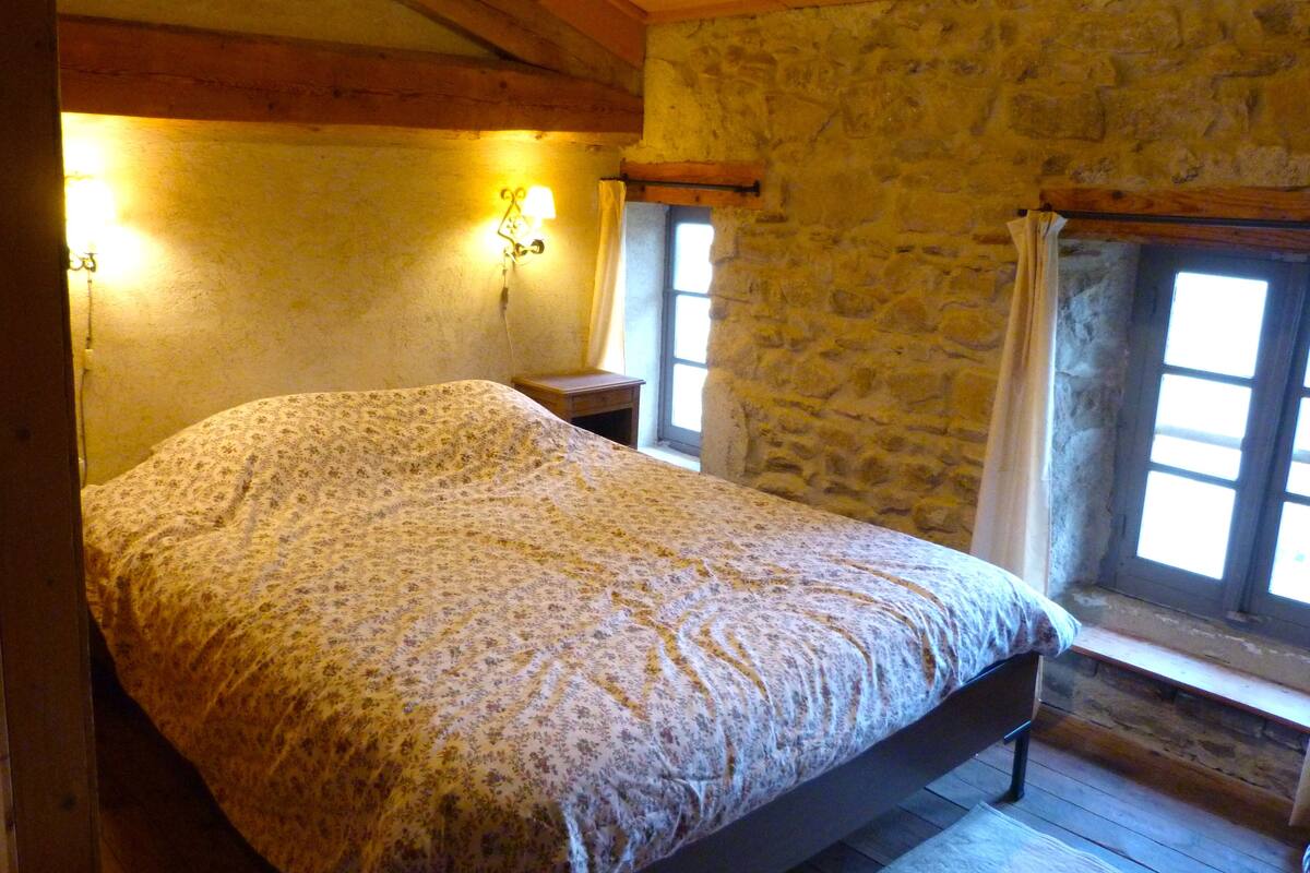Ein Schlafzimmer mit Holzbalkendecke, Steinwänden, einem Bett mit Blumenmuster-Bettwäsche, zwei Wandlampen und einem Fenster mit Vorhang.