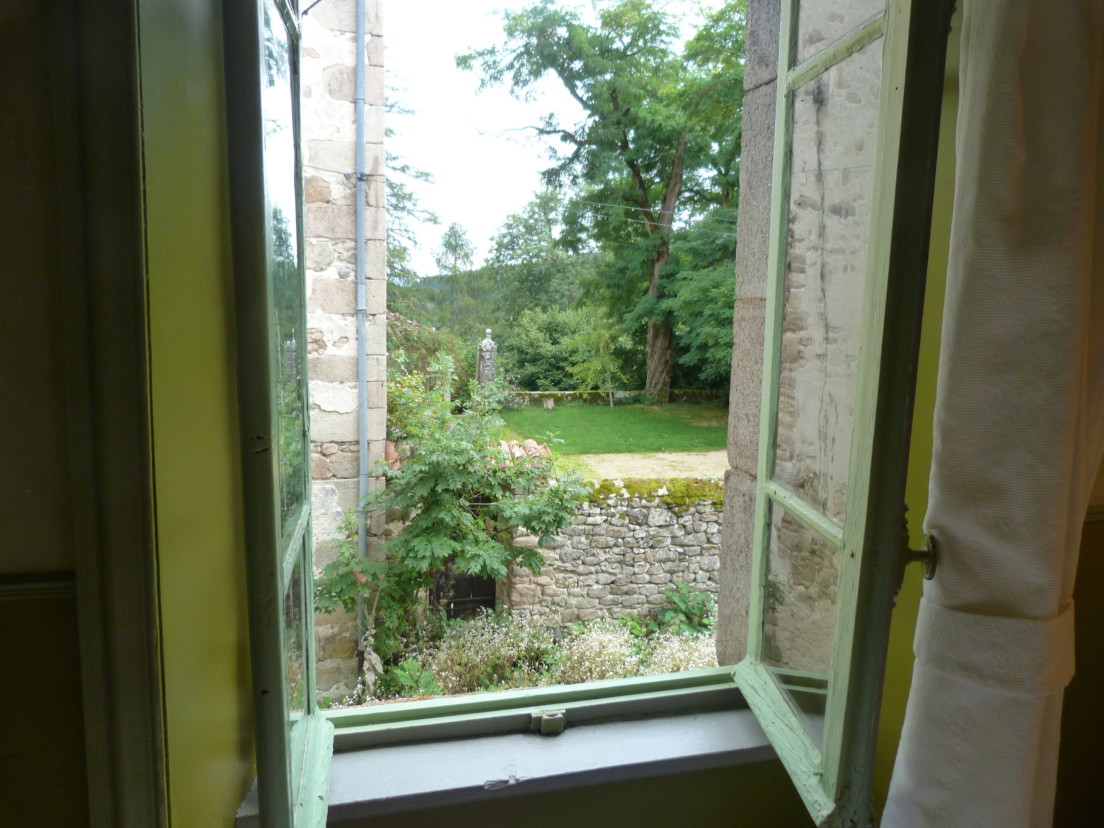 Une vue depuis une fenêtre, avec des murs en pierre et un espace verdoyant visible à l’extérieur.