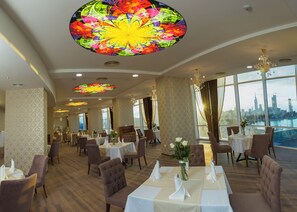 A spacious dining area with high ceilings, large windows, and a city view. - JRW Welmond Hotel (Batumi)