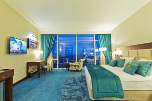 A hotel room with a large bed, a desk, a chair, a television, and a view of the cityscape. - JRW Welmond Hotel (Batumi)