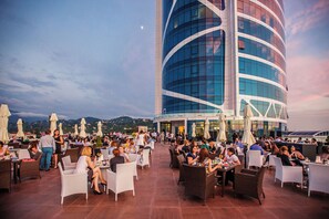 An outdoor dining area with tables and chairs, a large building with a curved facade, and a mountainous backdrop. - JRW Welmond Hotel (Batumi)
