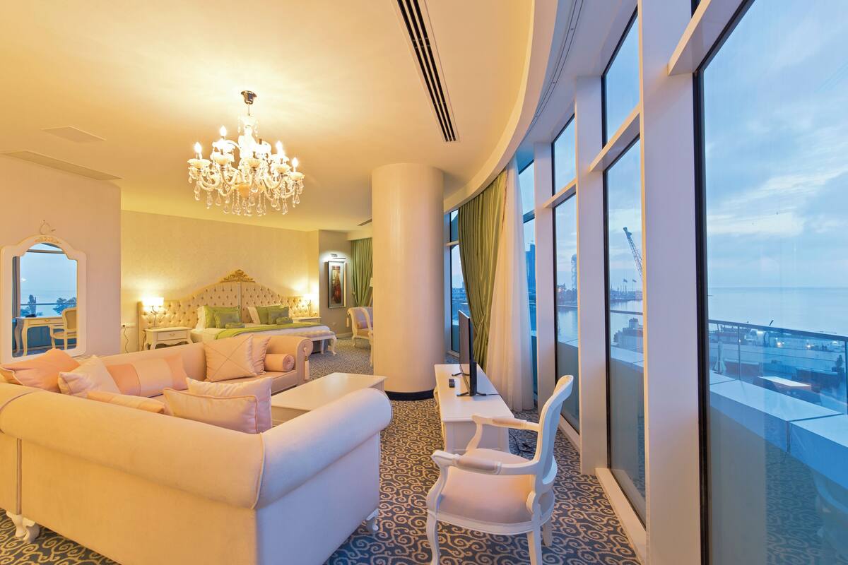 A hotel room with a large sofa, a dining table, a chandelier, and a view of the city.