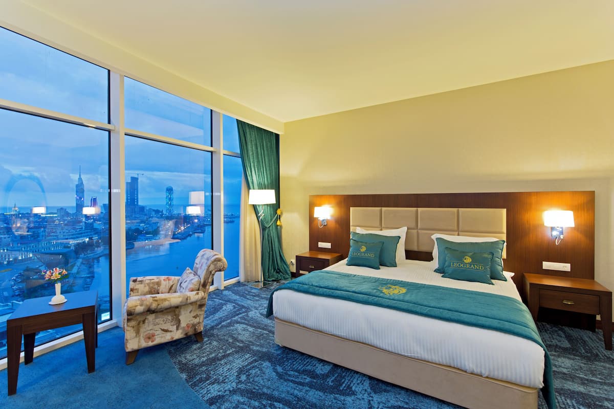 A hotel room with a large bed, two bedside tables, a chair, and a view of a cityscape through floor-to-ceiling windows.