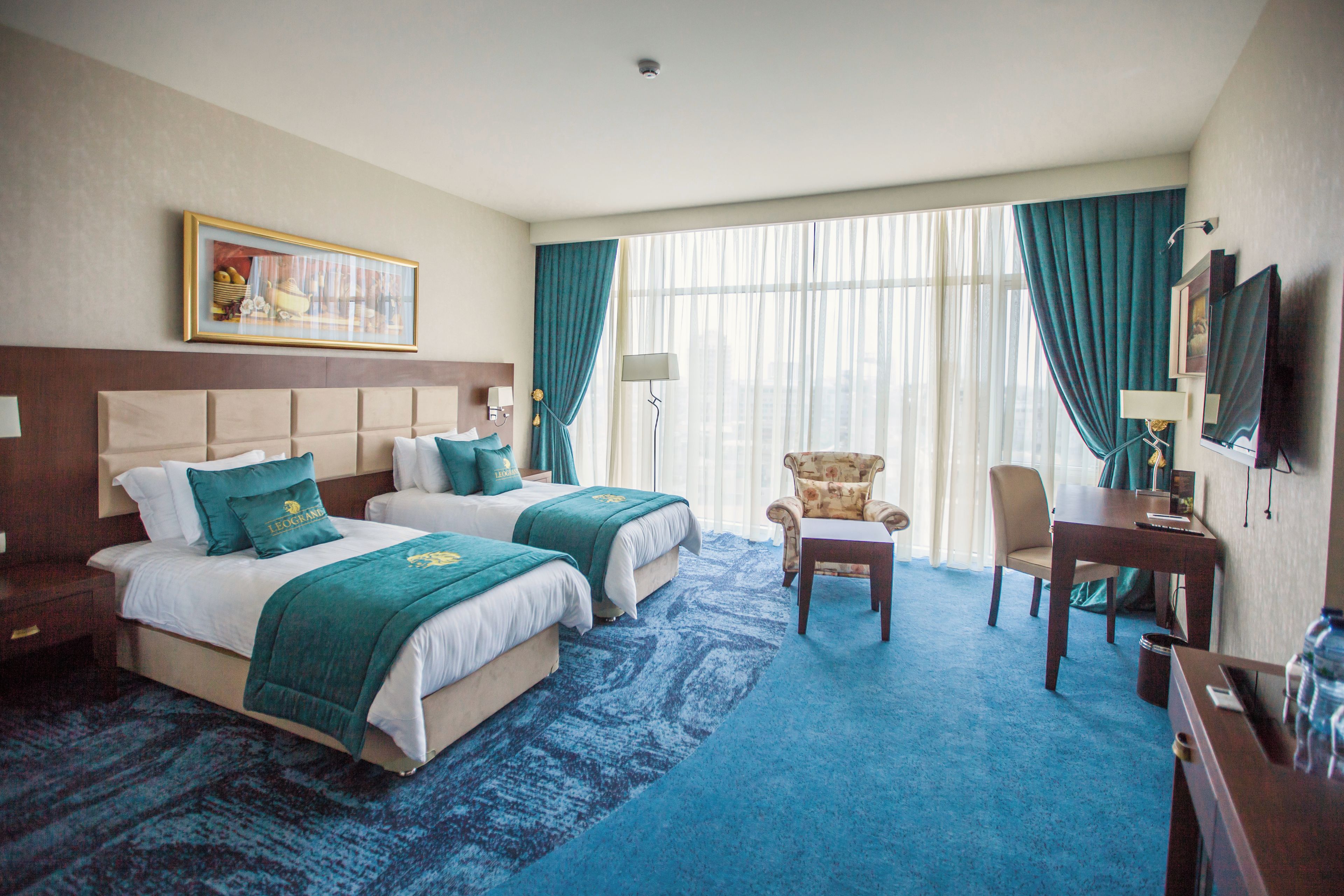 A hotel room with two beds, a desk, a chair, a lamp, a painting on the wall, and a large window with curtains.