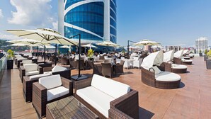 Un lounge en la azotea con mobiliario de mimbre, sombrillas y vistas a un edificio moderno.