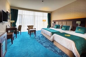 A hotel room with two beds, a desk, a chair, a TV, and a large window with curtains. - JRW Welmond Hotel (Batumi)