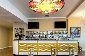 A bar area with a central bar counter, two bar stools, a decorative ceiling light, and a wall-mounted television. - JRW Welmond Hotel (Batumi)