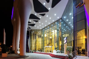 A modern building with a glass facade, a large entrance with steps, and a security camera on the right. - JRW Welmond Hotel (Batumi)