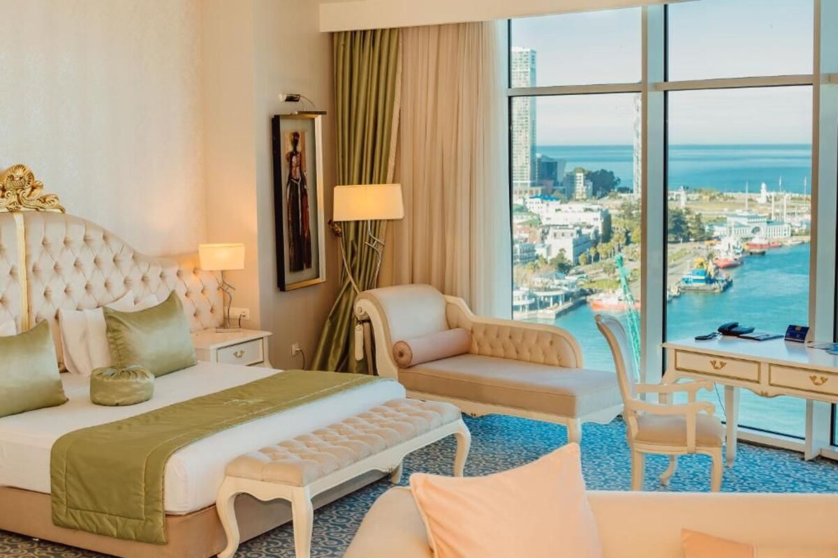 A hotel room with a large bed, a desk, a chair, and a view of a cityscape and water.