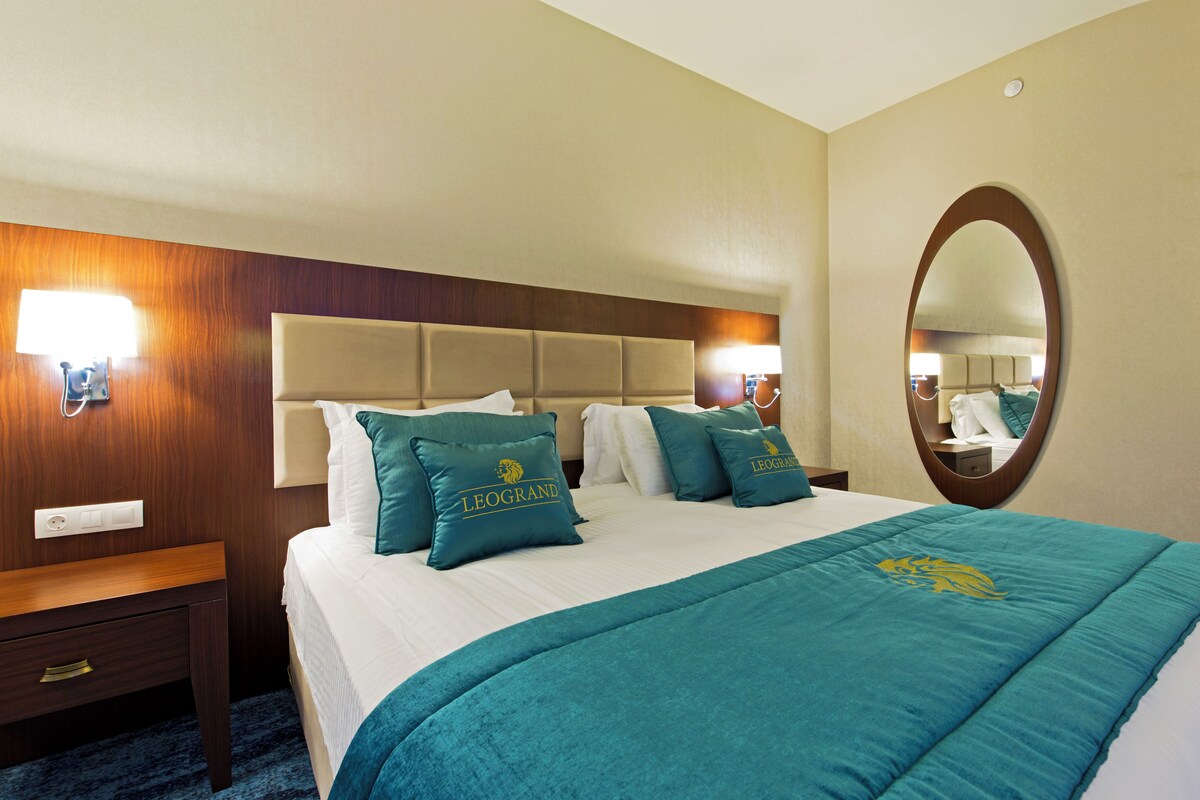A hotel room with a bed, a nightstand, a mirror, and a wall-mounted lamp.