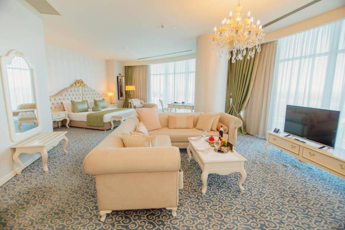 A hotel room with a large sofa, a bed with a headboard, a TV, and a chandelier.