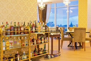 A bar area with a variety of liquor bottles, a cart with glasses, and a view of a cityscape through large windows. - JRW Welmond Hotel (Batumi)