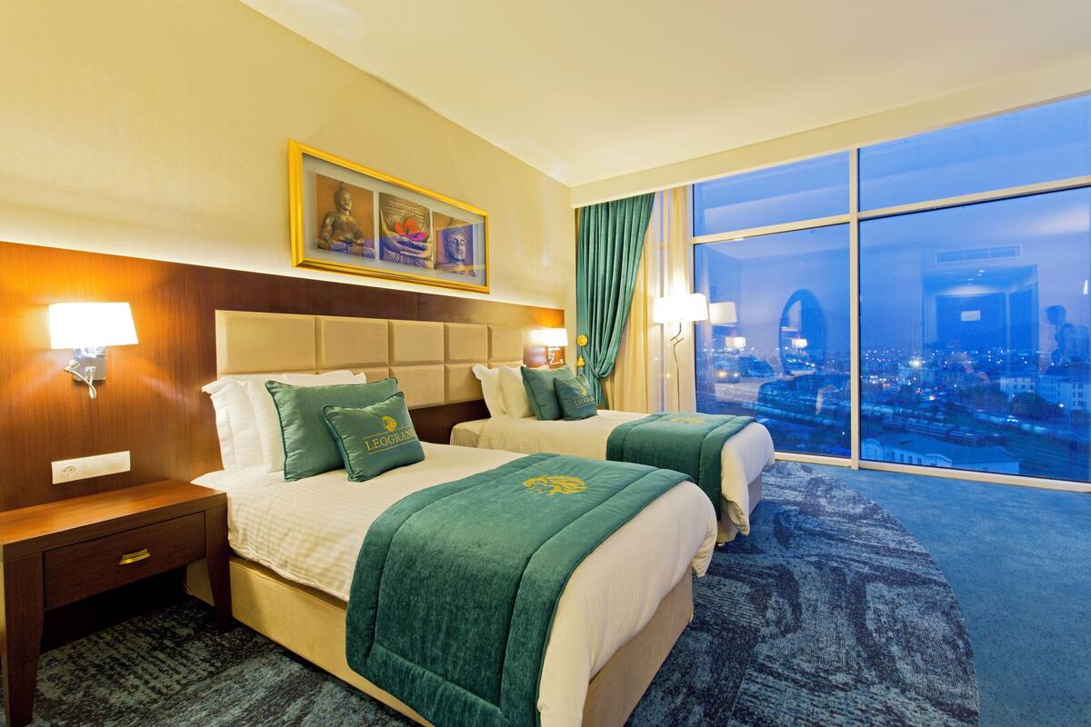 A hotel room with two beds, a headboard, a nightstand, a lamp, a framed picture, and a large window with a city view.