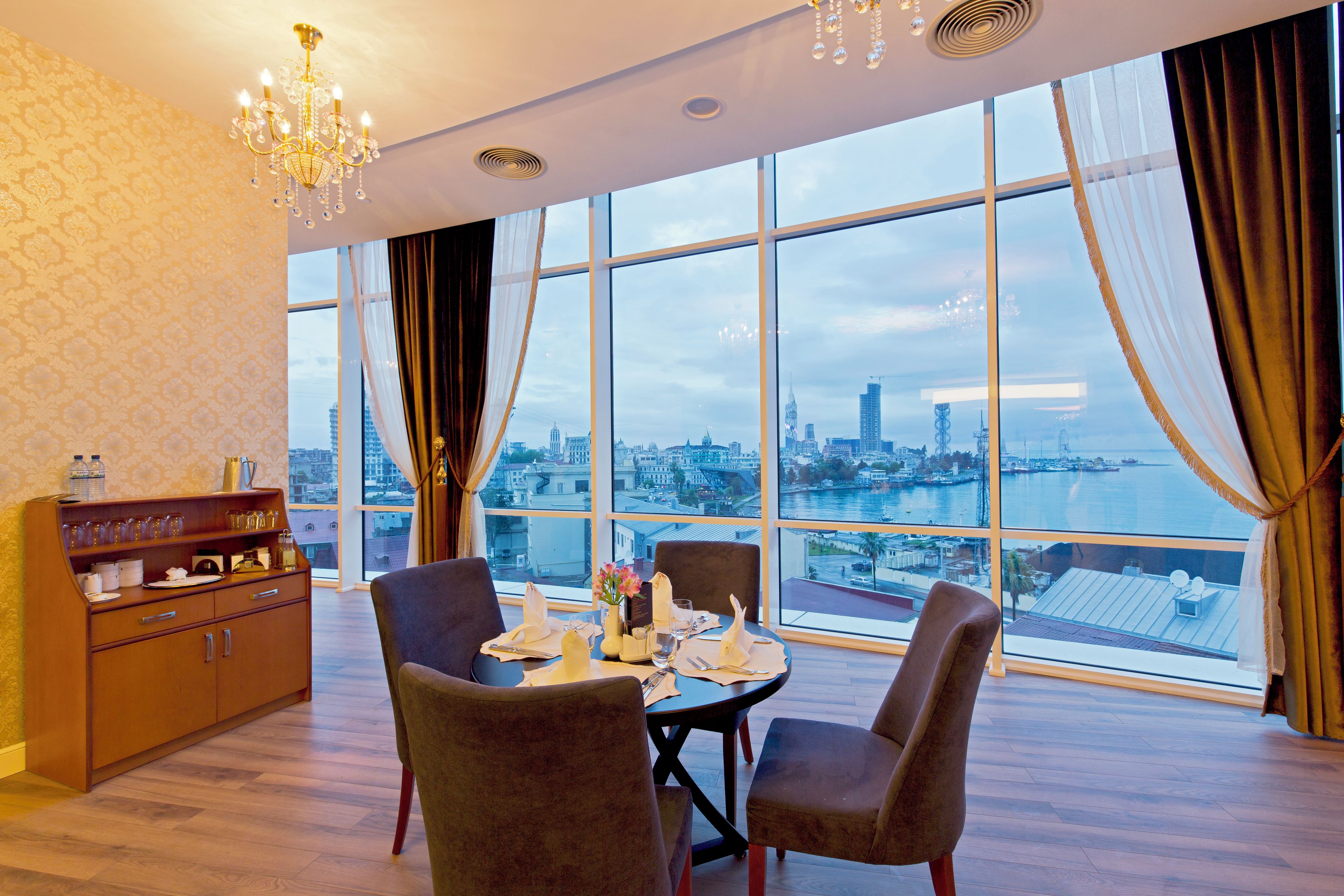 Comedor con mesa dispuesta para cuatro, candelabro y vista de un paisaje urbano a través de amplios ventanales.