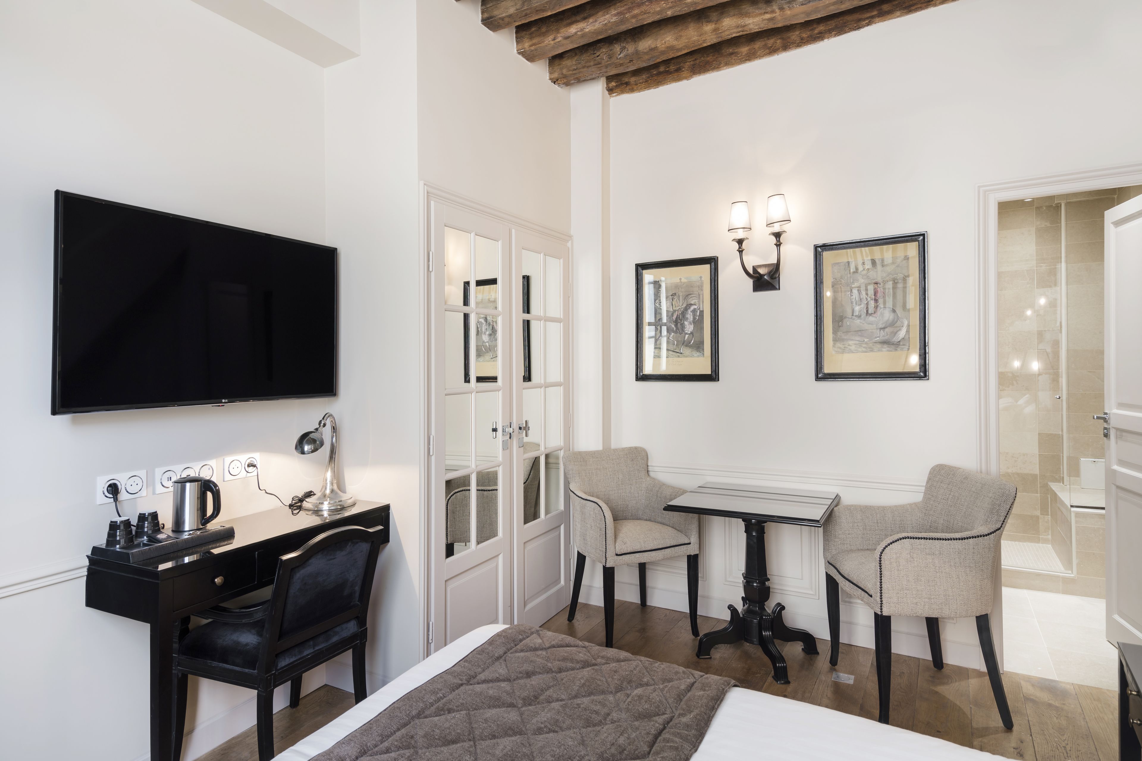 Habitación de hotel con cama, escritorio y silla, una mesa con dos sillas, televisión y obras de arte en las paredes.