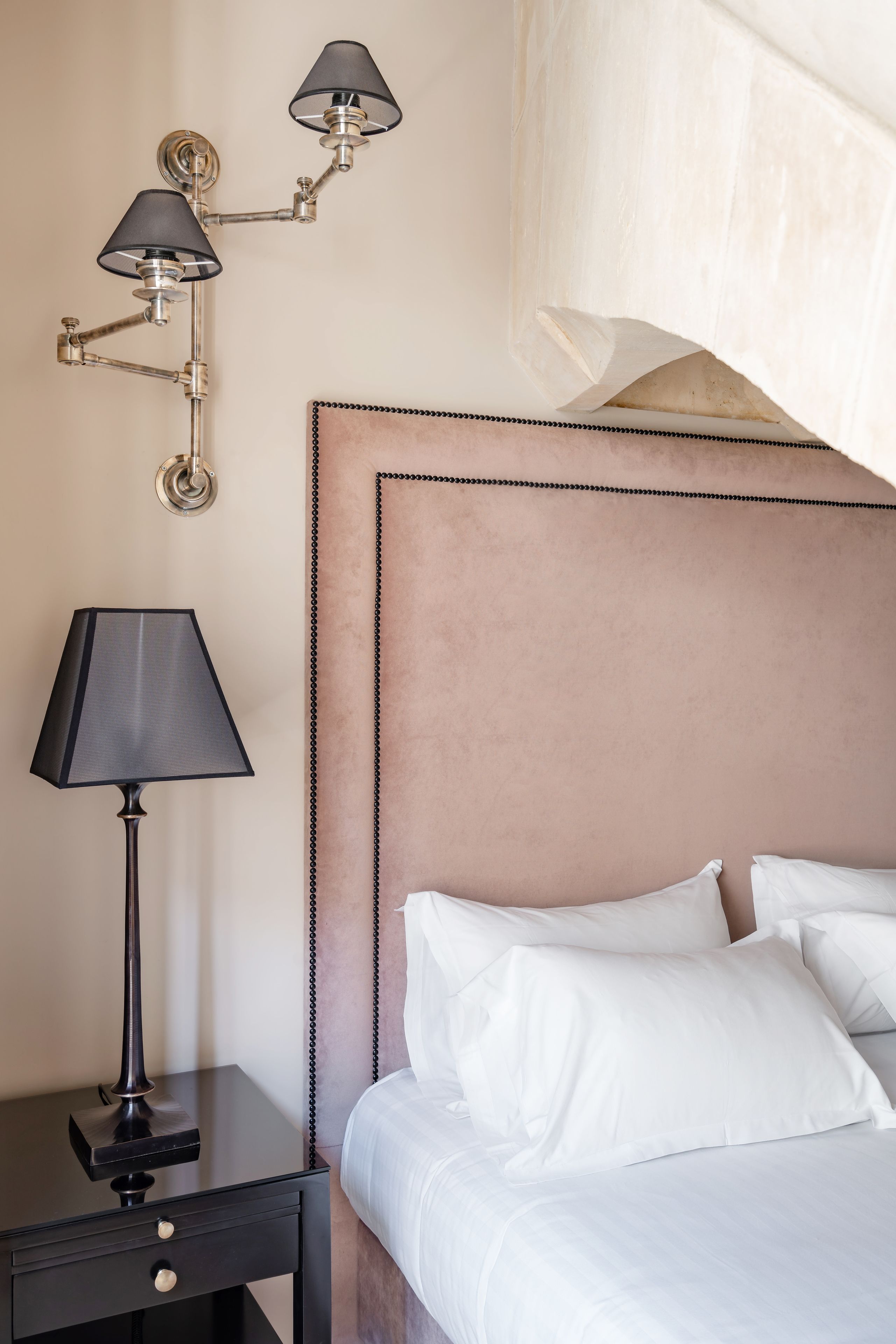 Una cama bien hecha con sábanas blancas, una mesita de noche negra con lámpara y un aplique de pared.