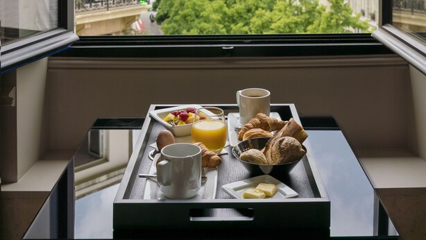 A breakfast tray with croissants, fruit, juice, and coffee on a window sill overlooking a cityscape with historic buildings and trees. - Hotel Le Presbytère (Paris)