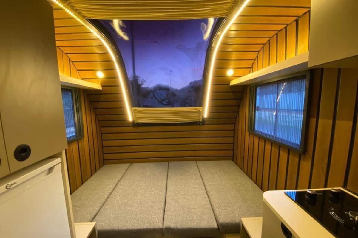 A compact RV interior with a bed, kitchenette, and a view of the night sky through a window.