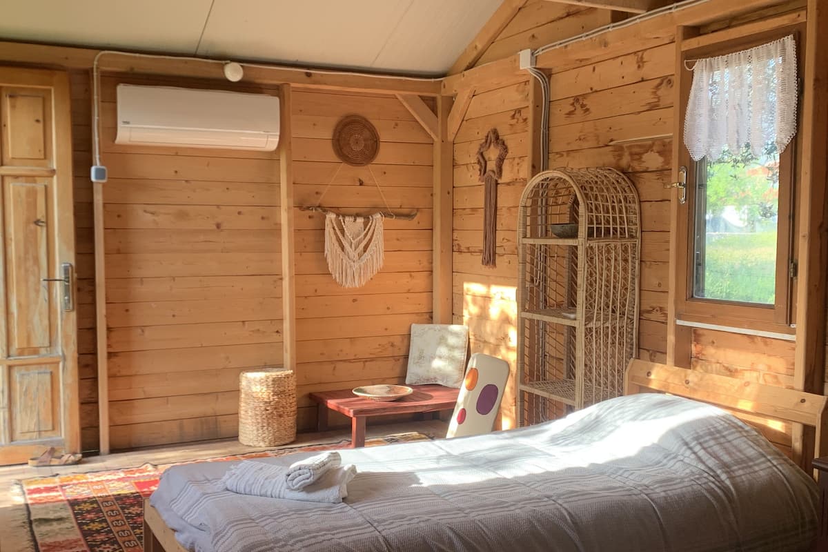 A cozy wooden cabin room with a bed, a small table, a woven basket, and a window with a view of greenery.