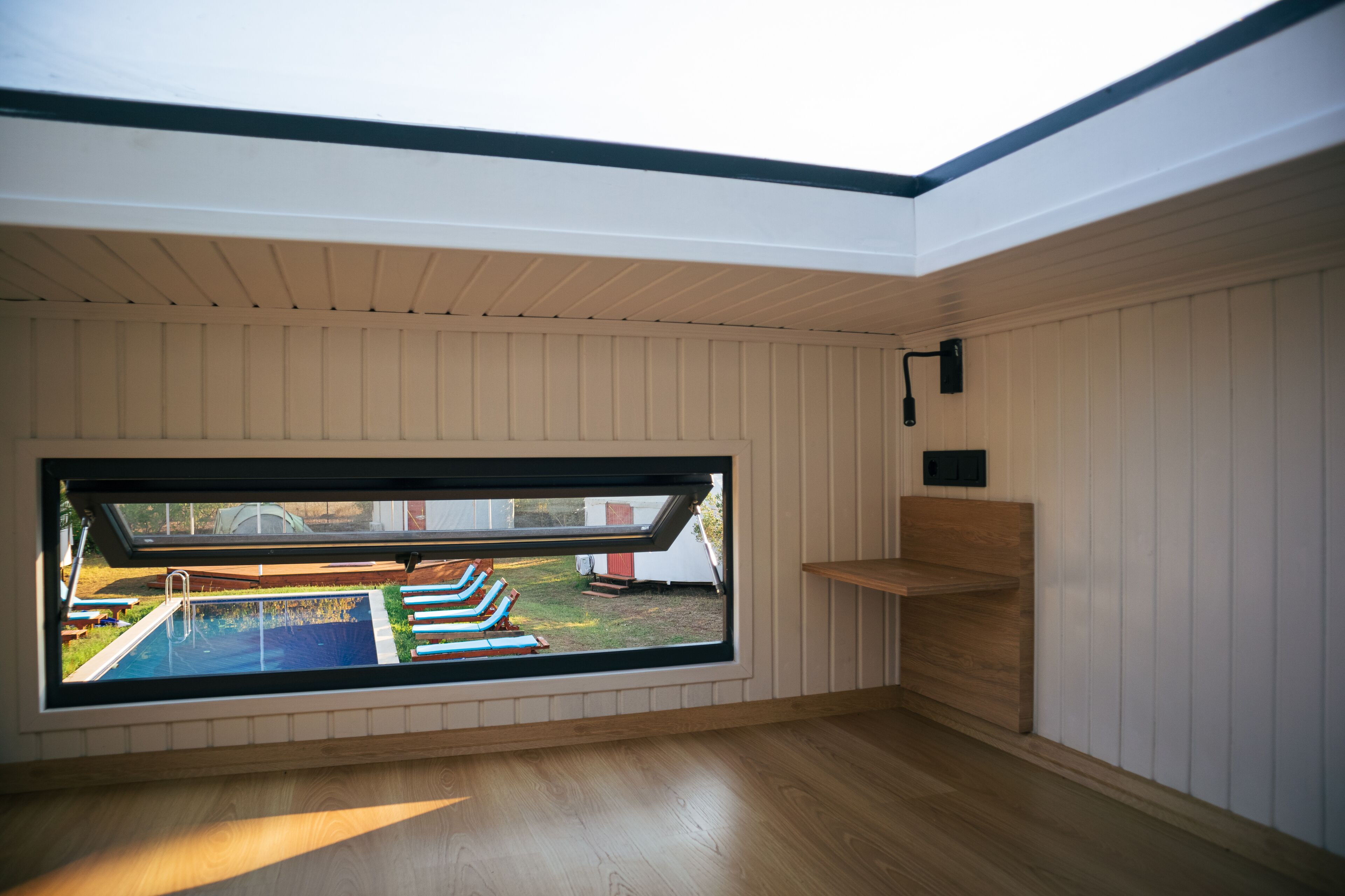 Ein Zimmer mit großem Dachfenster, Holzboden und Blick auf einen Swimmingpool und Gartenmöbel.