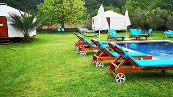 A pool area with lounge chairs and a tent, surrounded by a grassy lawn and trees, with a mountain in the background.