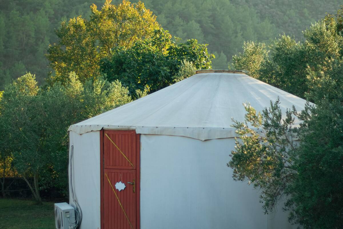 A white yurt with a red door, surrounded by greenery and trees.