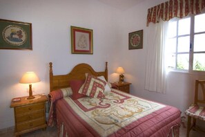 Un dormitorio con una cama de madera, dos mesitas de noche con lámparas, una silla y tres cuadros enmarcados en la pared.