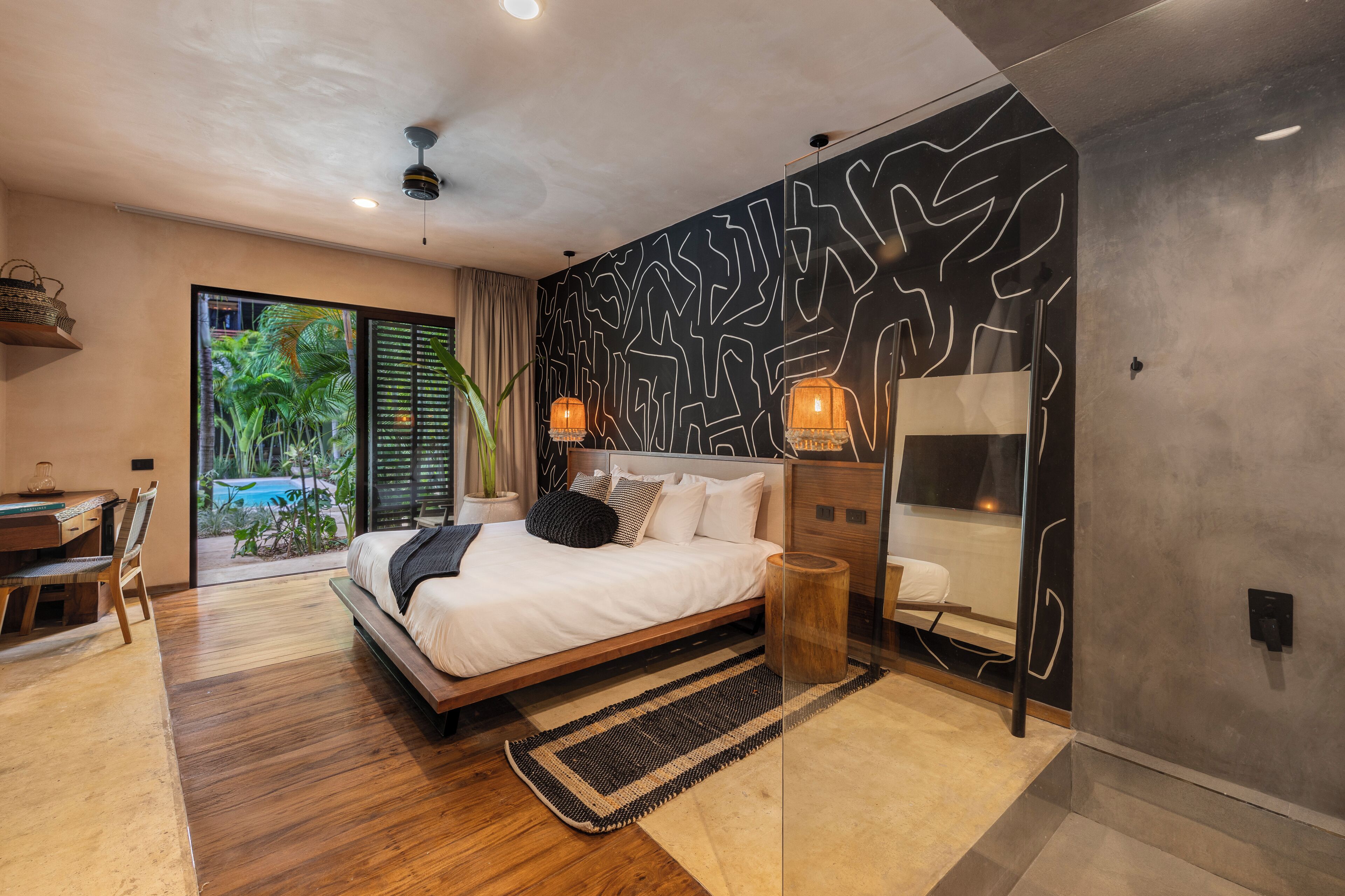 A modern bedroom with a large bed, a desk, and a view of a tropical garden.
