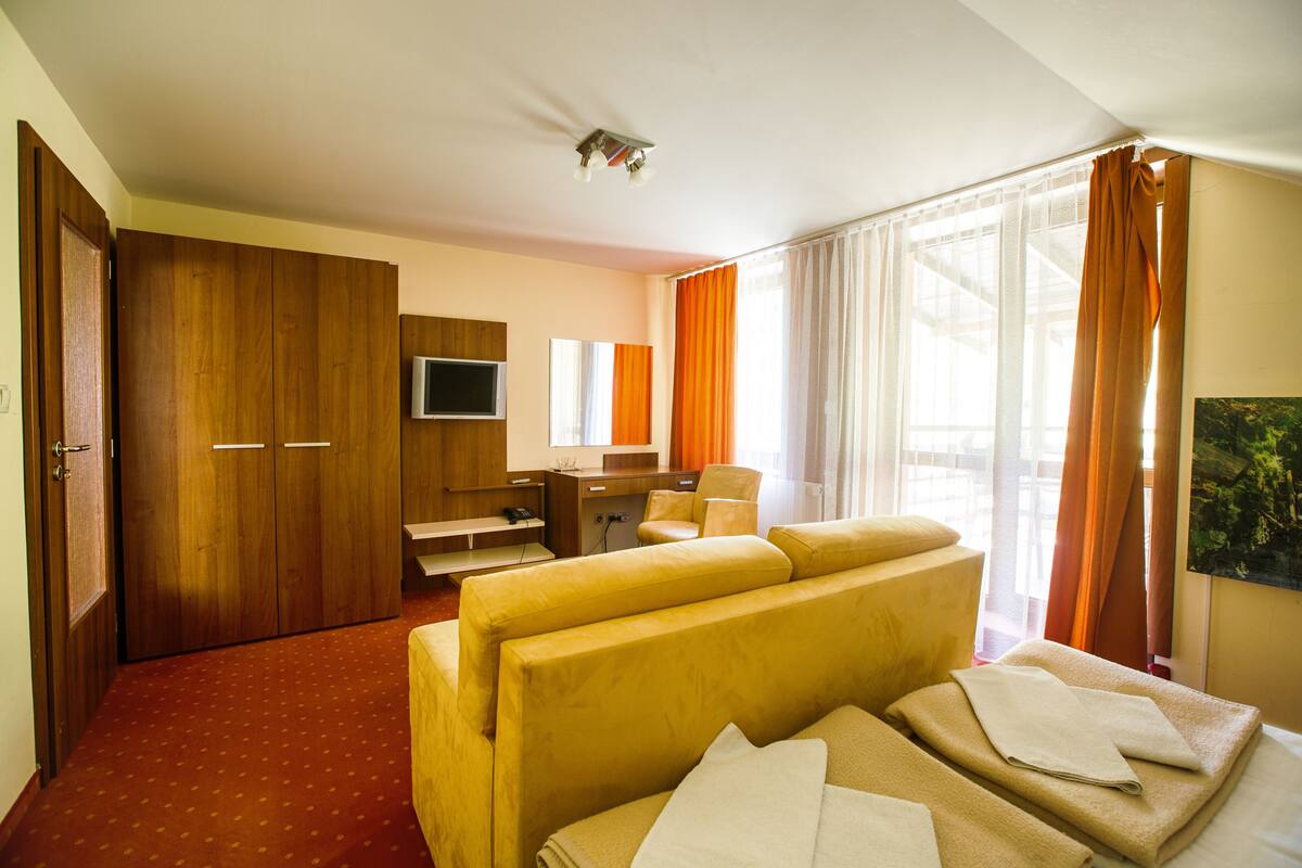 A hotel room with a sofa bed, a desk with a chair, a television, and a large window with curtains.
