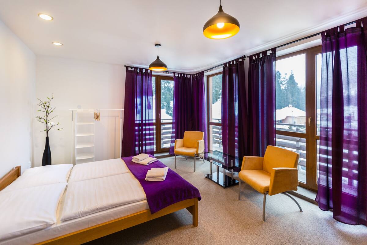 A hotel room with a bed, two chairs, a glass table, purple curtains, and a view of trees through large windows.