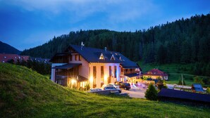 A large, multi-story building with a sloped roof, surrounded by a lush green landscape and a forested hill in the background. - Apartmány Mlynky (Mlynky)