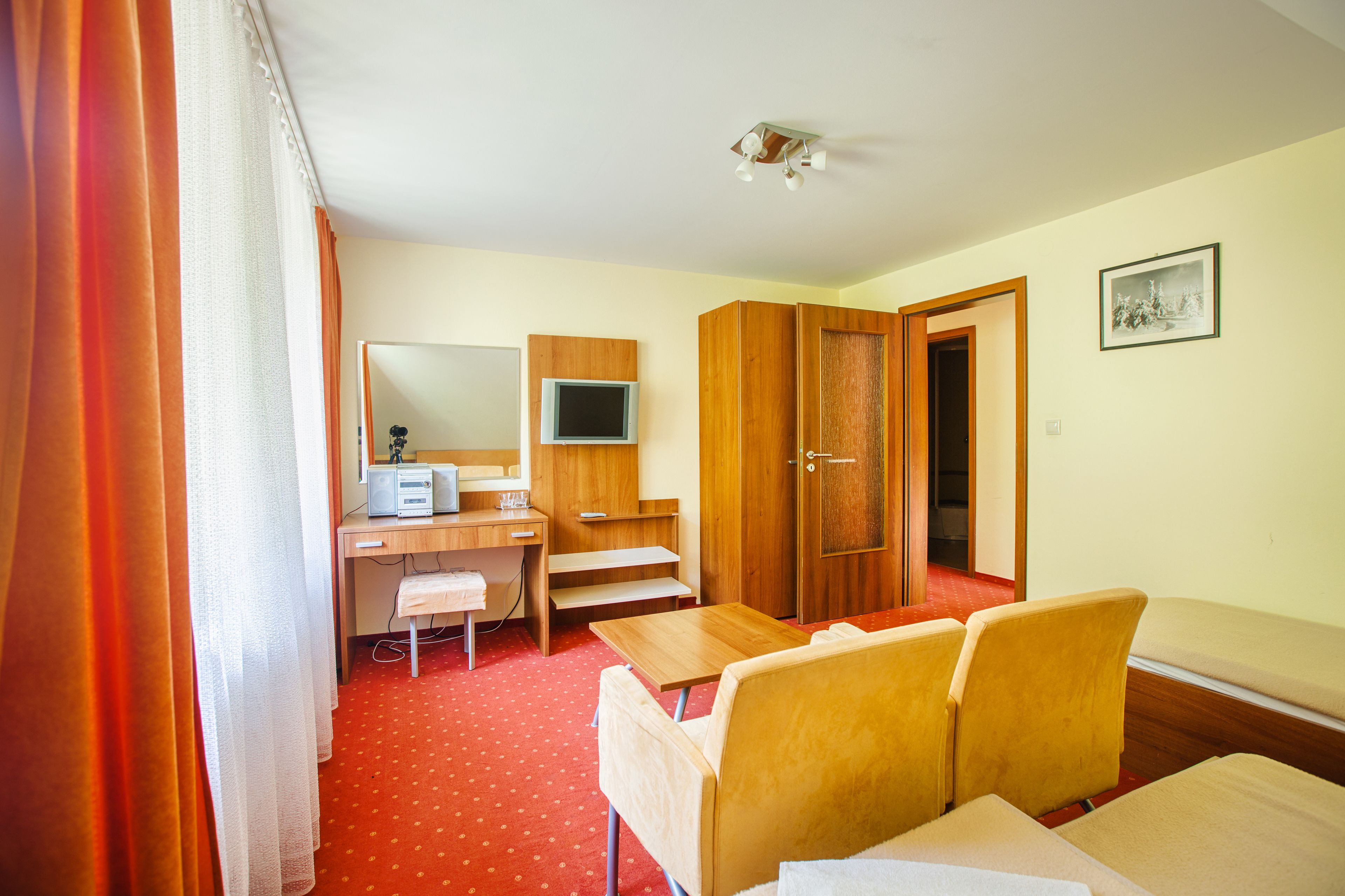 A hotel room with a desk, a television, a wardrobe, a bed, and a red carpet.