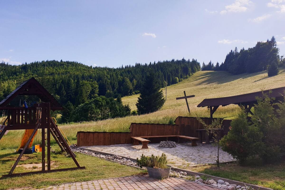 A playground with a slide and swings, picnic tables, a cross, and a small building with a sloped roof.