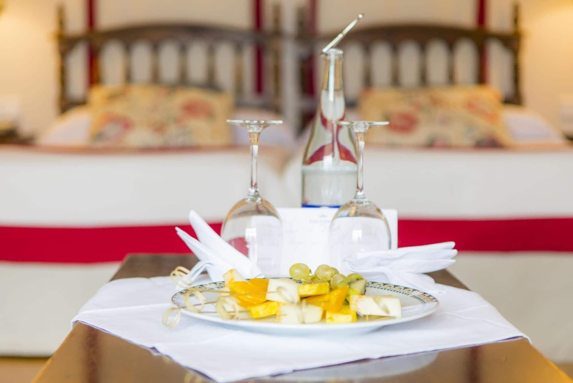 Una mesa de desayuno ordenada con un plato de fruta y dos copas de vino, frente a una cama con ropa de cama estampada en una habitación de hotel.