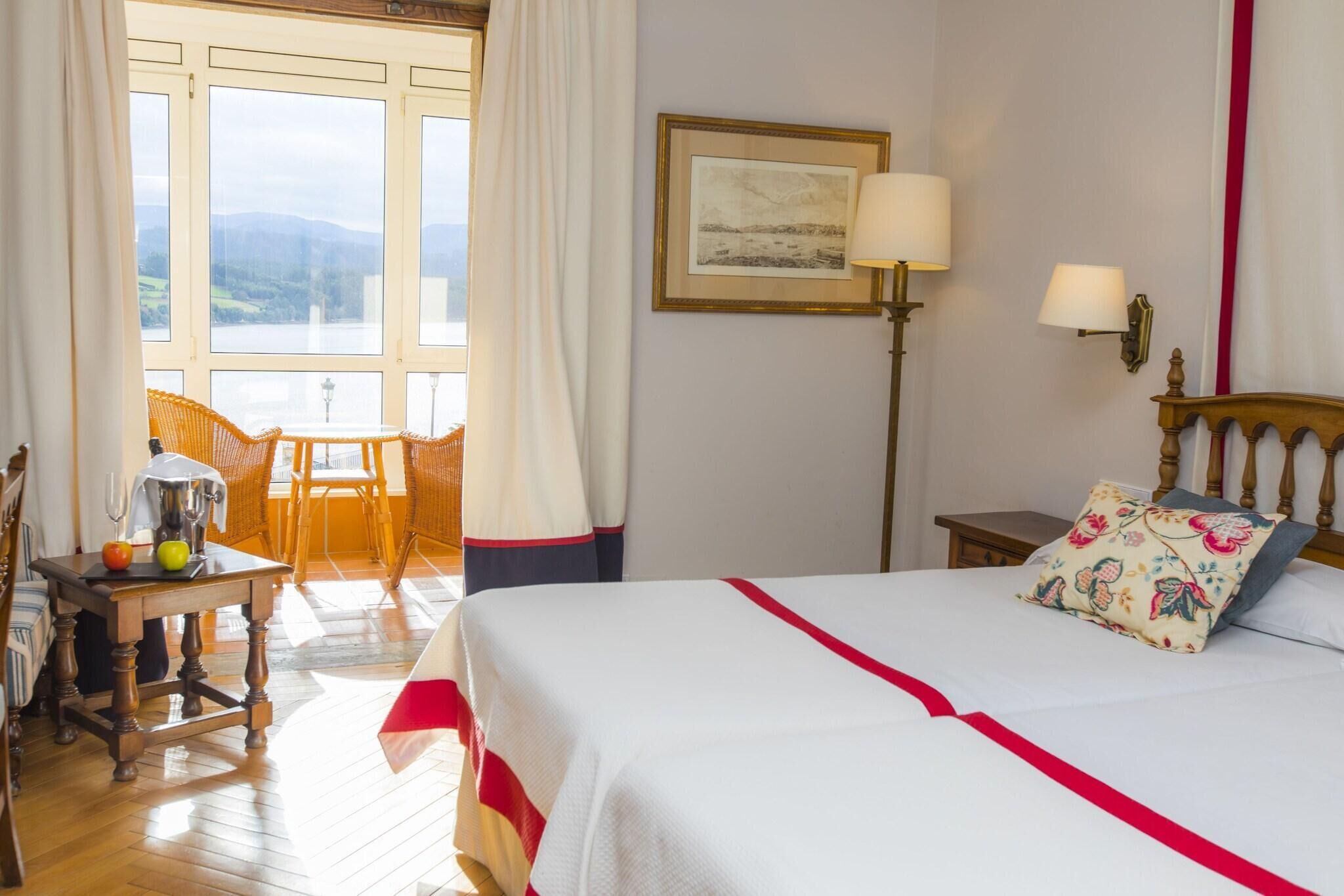 Ein Hotelzimmer mit Bett, einem Tisch mit Obst, einem Stuhl, einer Lampe und einem Fenster mit Blick auf die Landschaft.