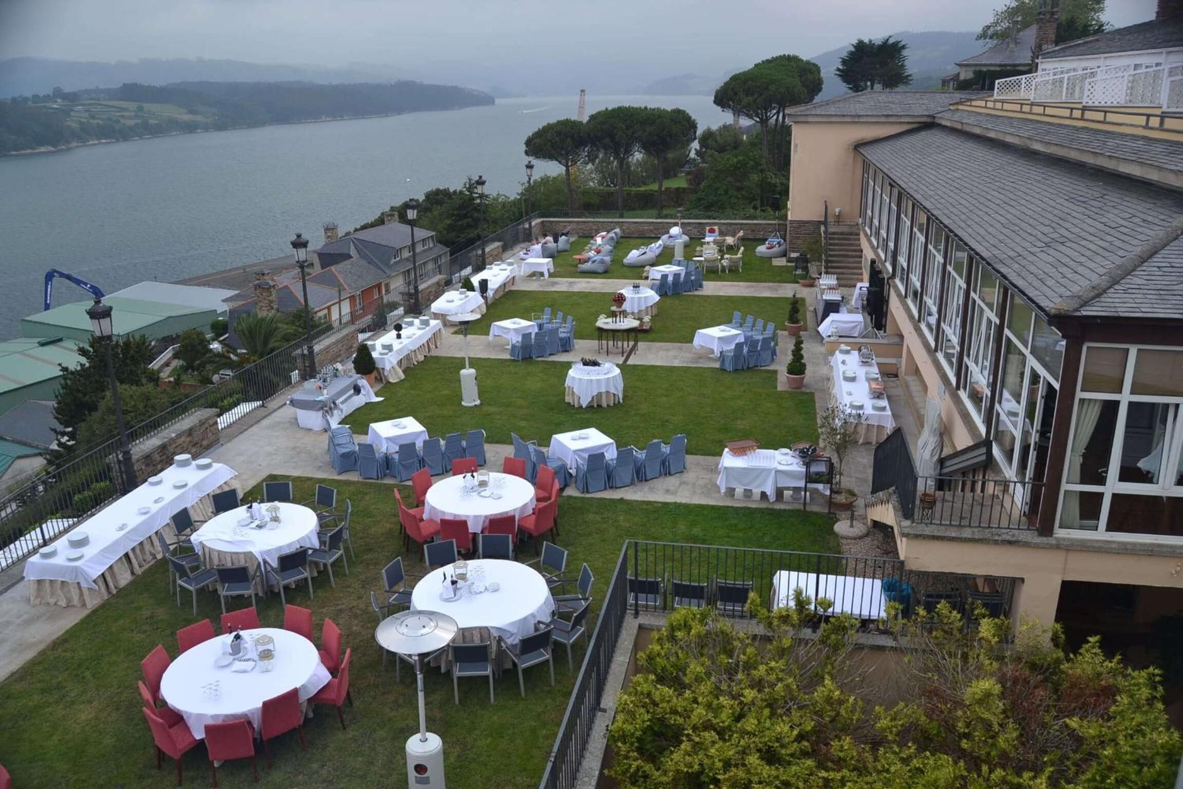An outdoor dining area with multiple round tables and chairs, set up on a grassy lawn with a view of a body of water and hills in the background.