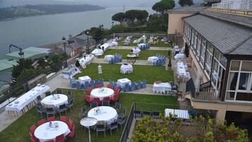 An outdoor dining area with multiple round tables and chairs, set up on a grassy lawn with a view of a body of water and hills in the background.