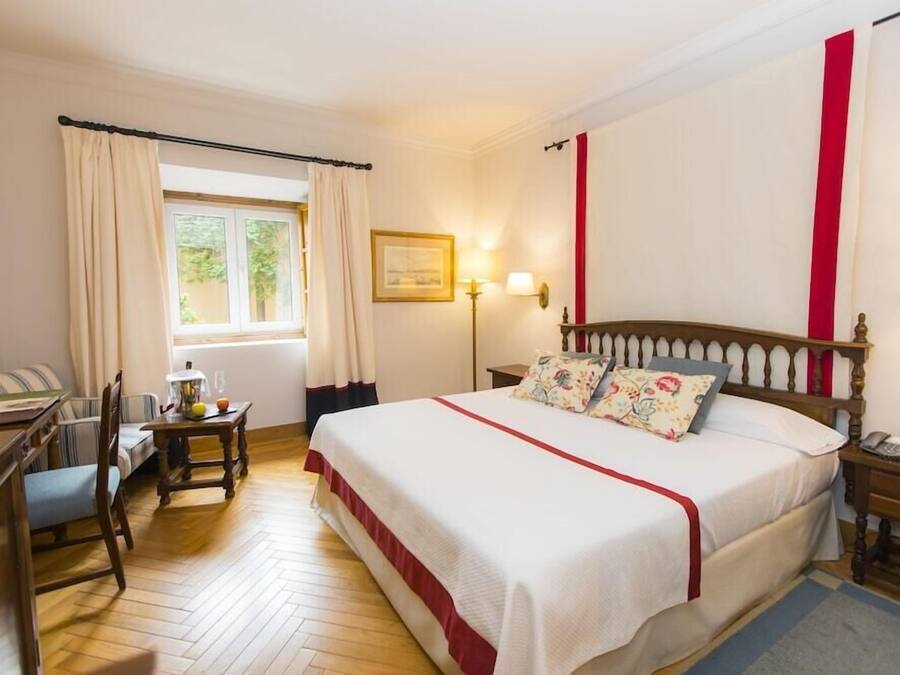 A hotel room with a large bed, a desk with a chair, a window with curtains, and a wall-mounted lamp.