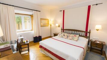 A hotel room with a large bed, wooden floor, a desk with a chair, a window with curtains, and a wall-mounted lamp.