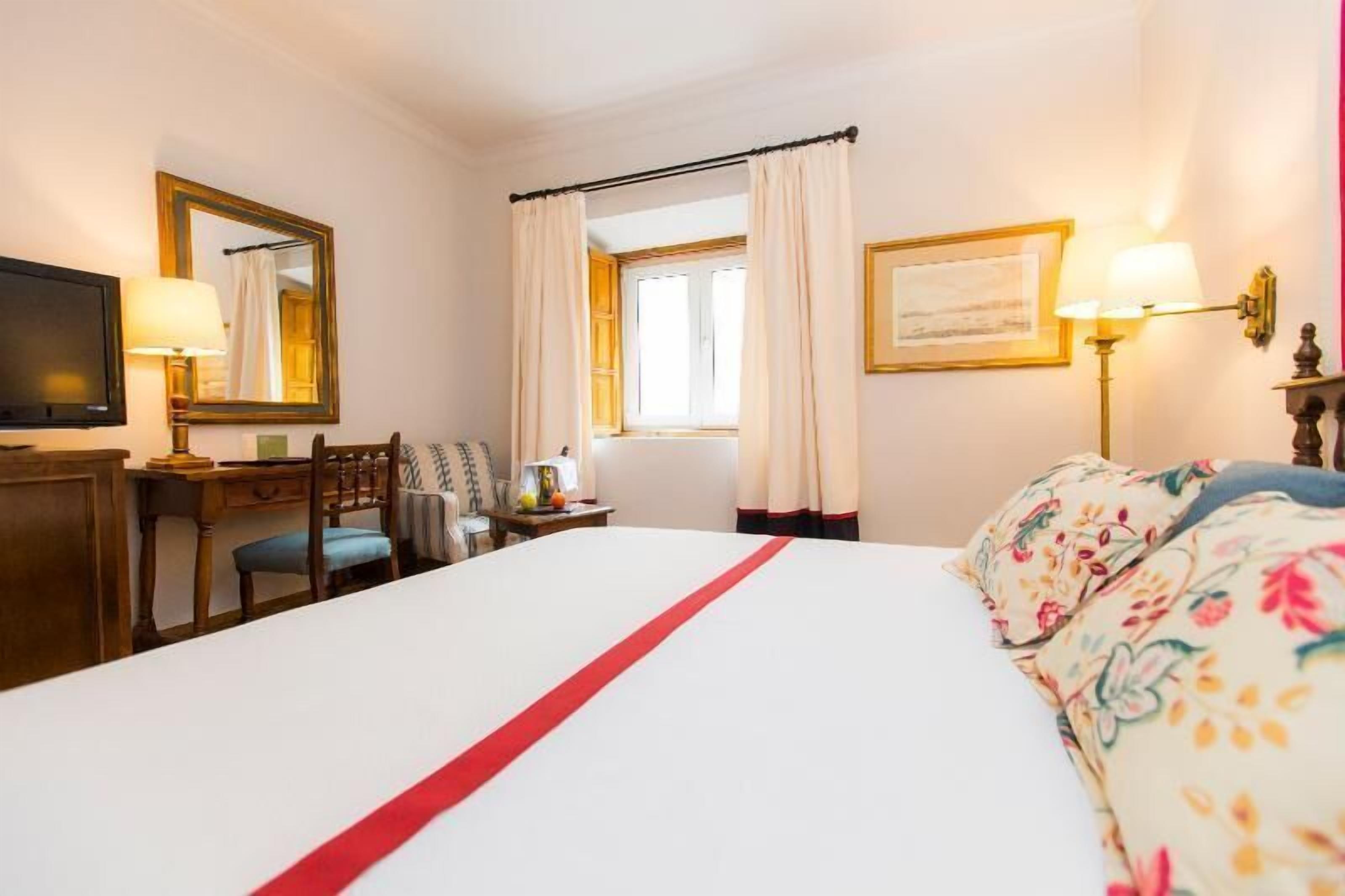 Una camera d'albergo con un letto, una scrivania, una sedia, un televisore e una finestra con tende.