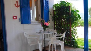 A small outdoor seating area with a table and four chairs, a vase with flowers, and a window with blue shutters. - White Rock Apartments (Samos)