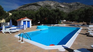 A swimming pool with a mountain backdrop, surrounded by lounge chairs and a small building with a red roof. - White Rock Apartments (Samos)
