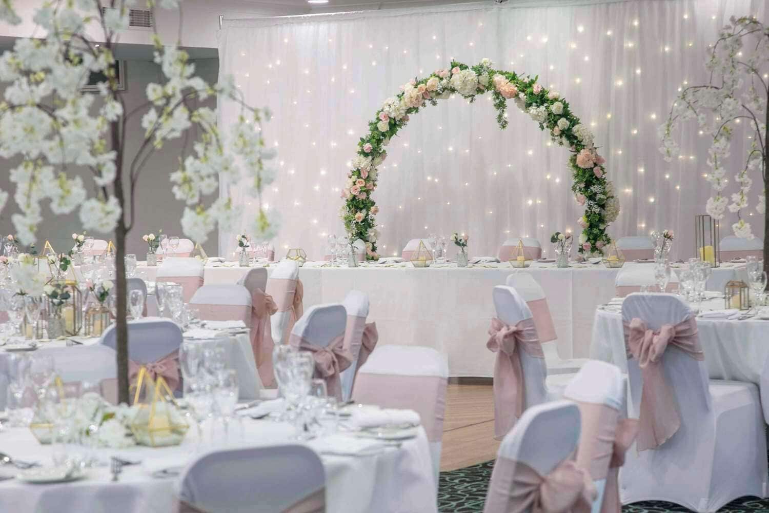 A banquet hall with tables set for a formal event, adorned with floral centerpieces and white drapery.