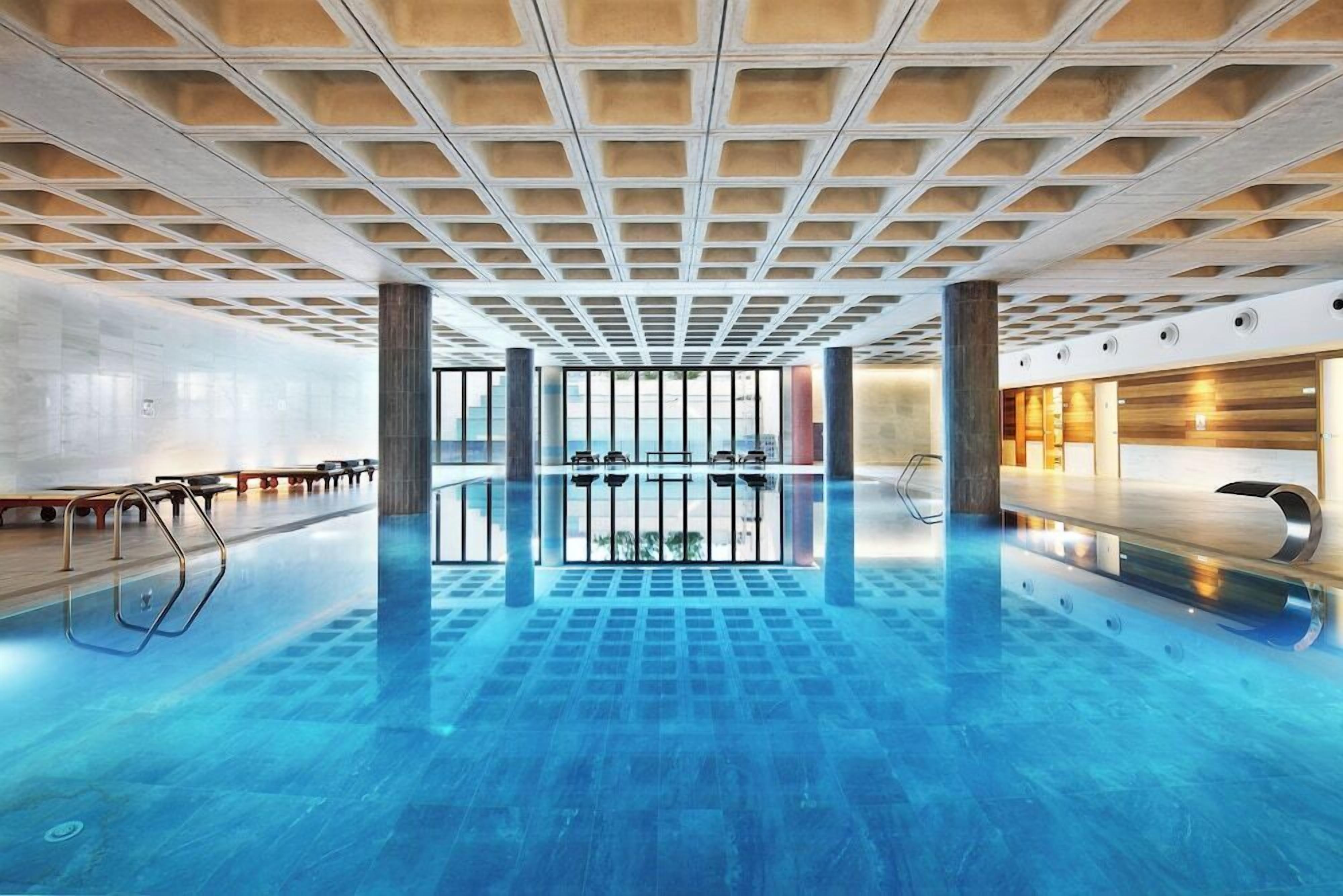 Piscina interior moderna con diseño geométrico en el techo, barandillas de vidrio transparente y superficie de agua limpia y reflectante.