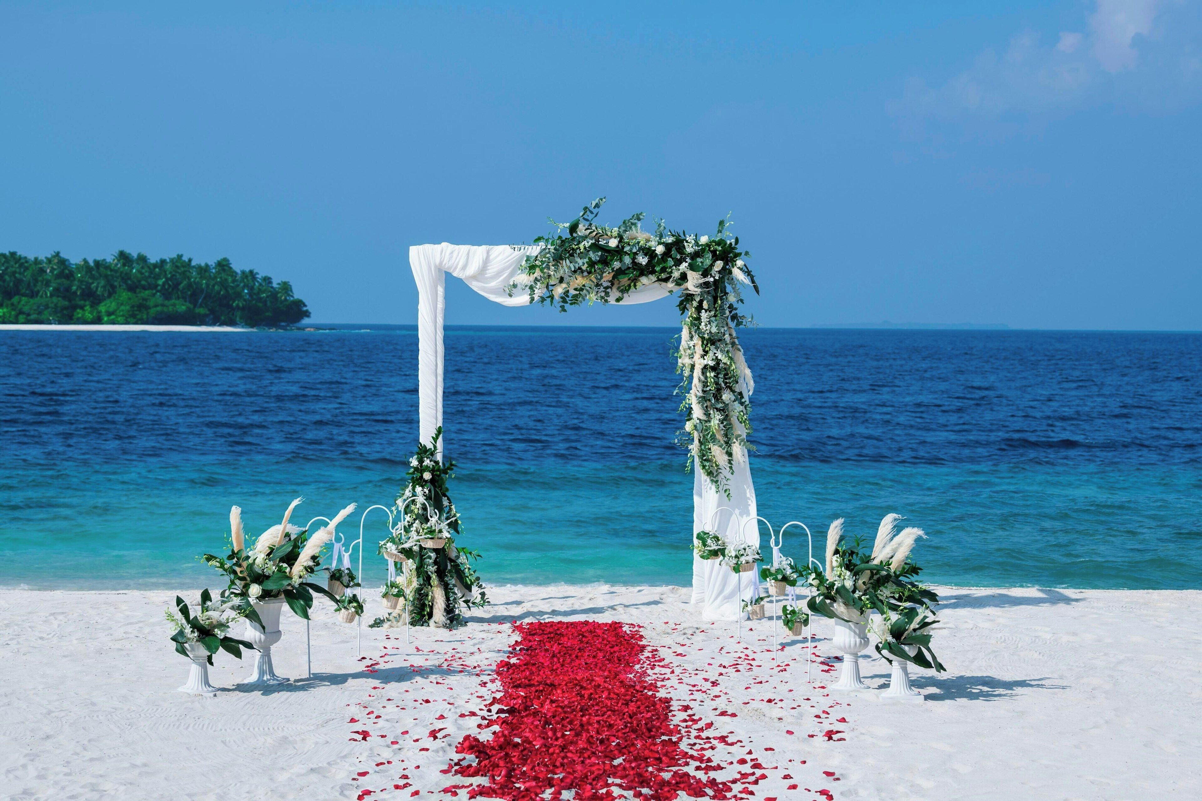 A beach wedding arch with white drapery and greenery, surrounded by white chairs with greenery, and a red flowered aisle on a sandy beach with a clear blue sea and a distant island.