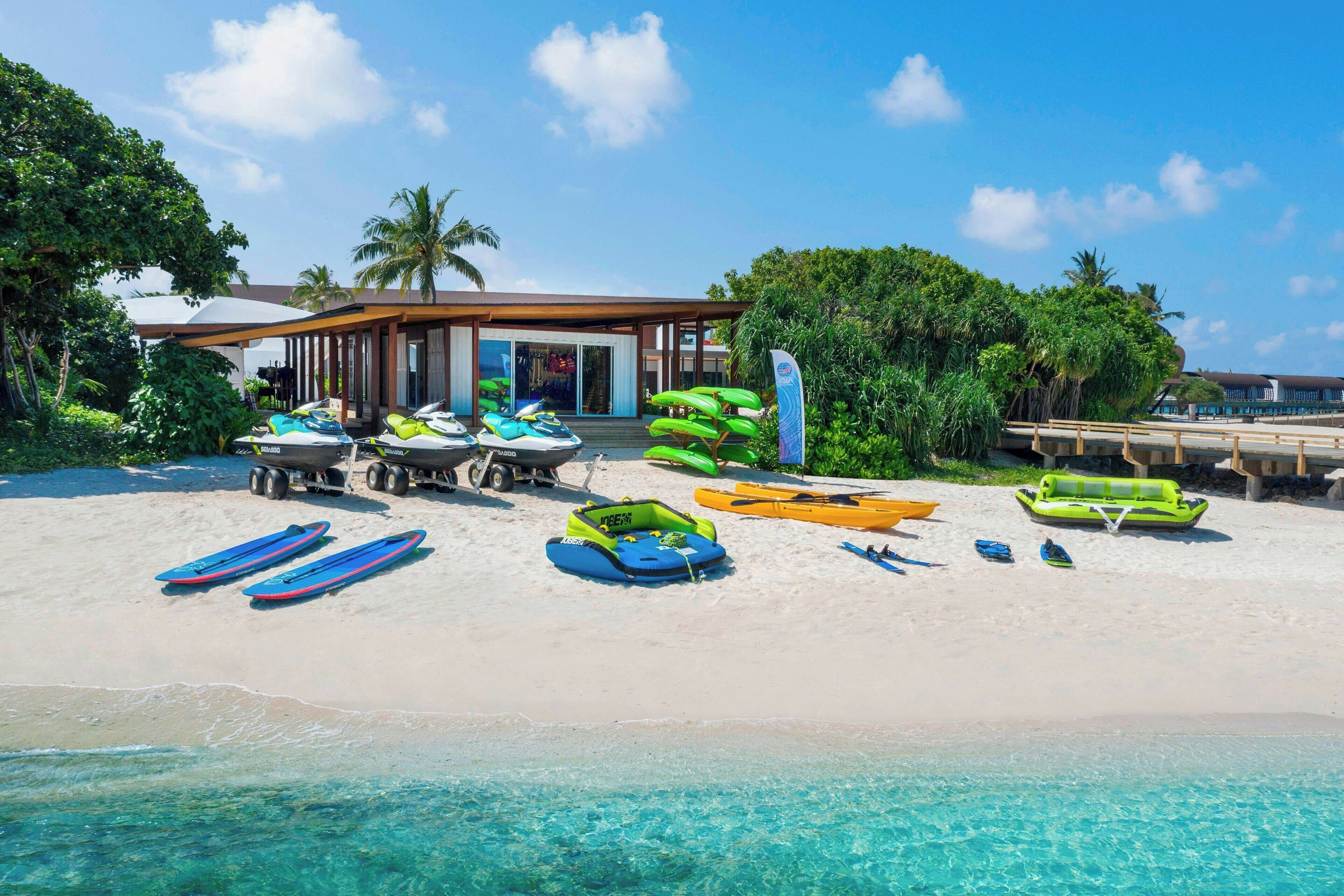 A beach with a building, clear turquoise water, and various watercraft.