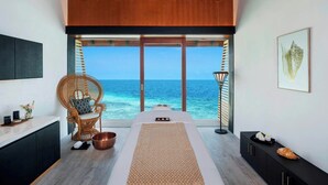 A massage room with a view of the ocean, a wicker chair, a massage table, and a black cabinet with a shell on top. - The Westin Maldives Miriandhoo Resort (Miriandhoo Island)